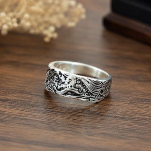 May include: A silver-toned metal ring featuring an intricate, embossed design of swirling waves and floral patterns. The band has a polished interior and a darkened, recessed finish within the relief details to highlight the textured surface.