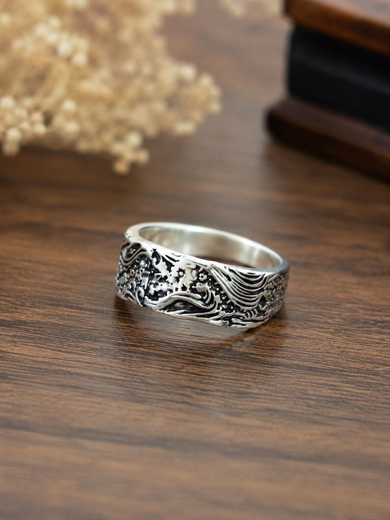 May include: A silver-toned metal ring featuring an intricate, embossed design of swirling waves and floral patterns. The band has a polished interior and a darkened, recessed finish within the relief details to highlight the textured surface.