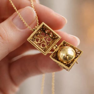 Puede incluir: Un collar con colgante dorado con un relicario calado cuadrado y abierto. El relicario está abierto, revelando una esfera dorada. La cadena y el relicario están sostenidos por una mano. El relicario tiene un diseño calado ornamental.