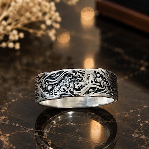 May include: A wide silver band ring featuring an intricate, raised relief pattern of swirling ocean waves and small floral elements. The metal has a polished finish with darkened recessed areas that highlight the detailed, textured design.