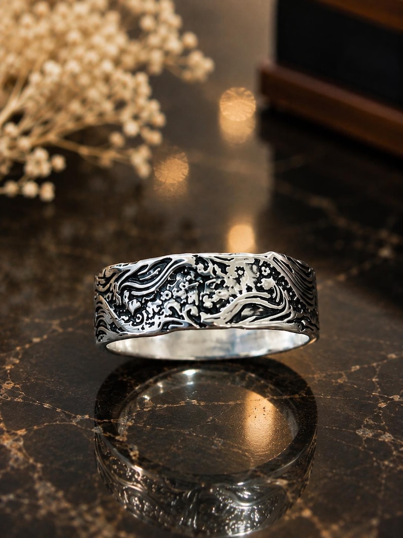 May include: A wide silver band ring featuring an intricate, raised relief pattern of swirling ocean waves and small floral elements. The metal has a polished finish with darkened recessed areas that highlight the detailed, textured design.