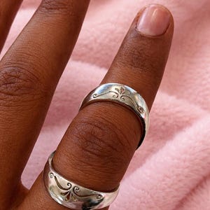 Könnte beinhalten: Zwei silberne Ringe mit einem dekorativen floralen Design werden auf einem Finger gezeigt. Die Ringe haben eine polierte Oberfläche und eine gebogene Form. Der Hintergrund ist ein weicher rosa Stoff.