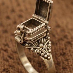 May include: A silver ring with an open, hinged compartment. The ring features ornate floral and beaded detailing on the band and around the compartment. The ring is set against a brown background.