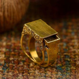 May include: A gold-colored ring with a rectangular top and a small drawer that slides out. The ring has ornate detailing on the sides and a double band. The ring is shown on a textured, multi-colored surface.