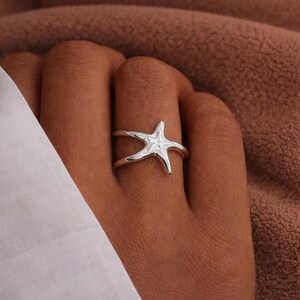 Puede incluir: Un anillo de plata con forma de estrella de mar. El anillo presenta un diseño de estrella de mar en la parte superior, con una banda que rodea el dedo. El anillo se lleva en una mano con un fondo marrón.