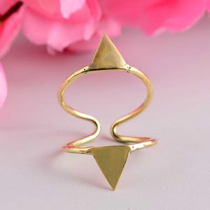 May include: A gold-colored ring with two triangular shapes, one at the top and one at the bottom, connected by a curved band. The ring has a minimalist design and is set against a soft pink background.