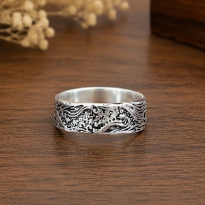 May include: A silver-toned band ring featuring an intricate, embossed relief pattern of swirling waves and small floral elements. The metal surface has a polished interior and a darkened, oxidized finish within the recessed areas of the design.