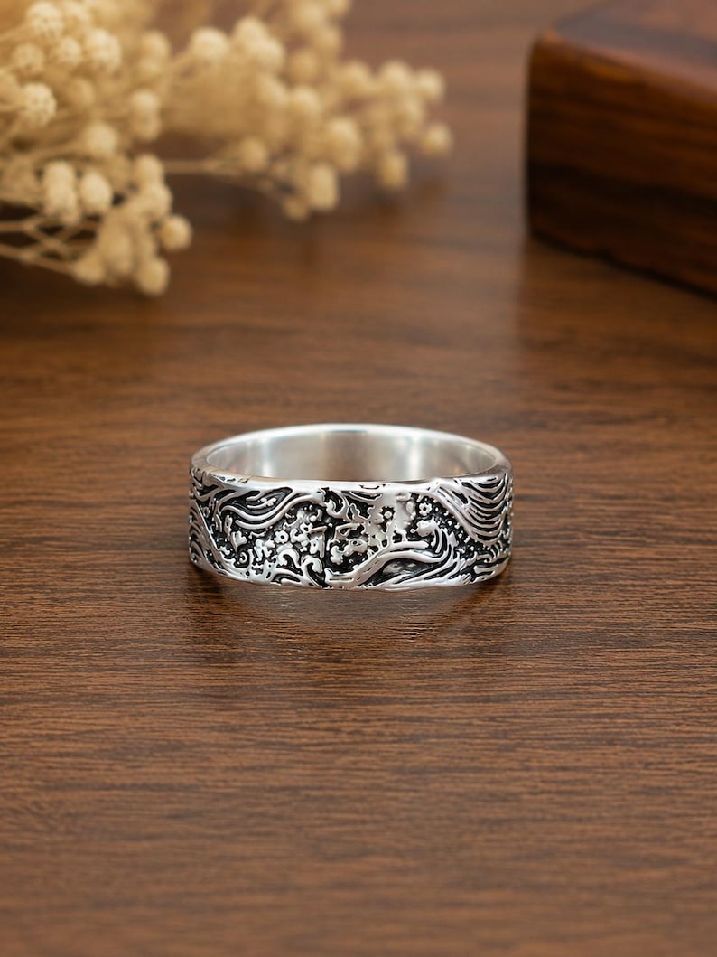 May include: A silver-toned band ring featuring an intricate, embossed relief pattern of swirling waves and small floral elements. The metal surface has a polished interior and a darkened, oxidized finish within the recessed areas of the design.