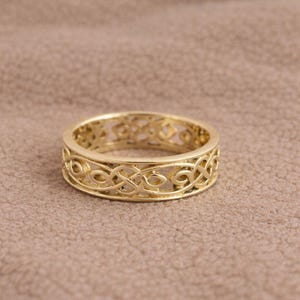 May include: A gold-colored ring with an intricate Celtic knot design. The ring has a band with a repeating pattern of interwoven lines and curves. The ring is set against a neutral background.