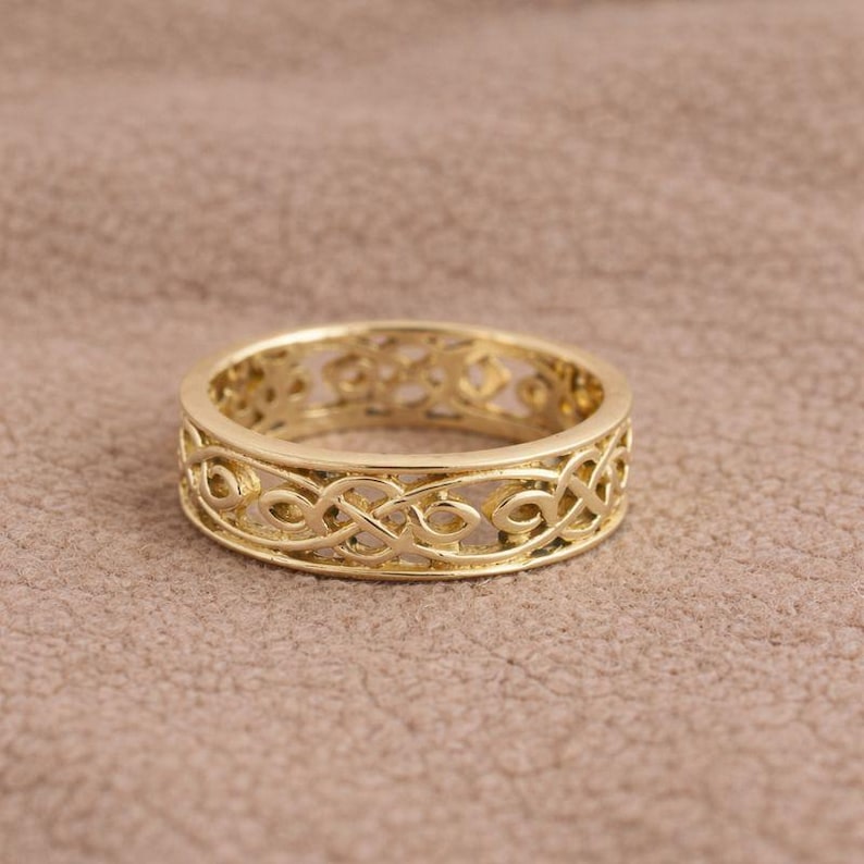 May include: A gold-colored ring with an intricate Celtic knot design. The ring has a band with a repeating pattern of interwoven lines and curves. The ring is set against a neutral background.