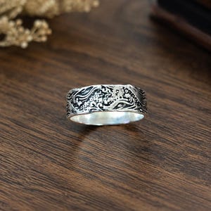 May include: A silver band ring featuring an intricate, raised relief pattern of swirling waves and small floral elements. The metal has a polished finish on the interior and a darkened, oxidized finish within the recessed areas of the design.