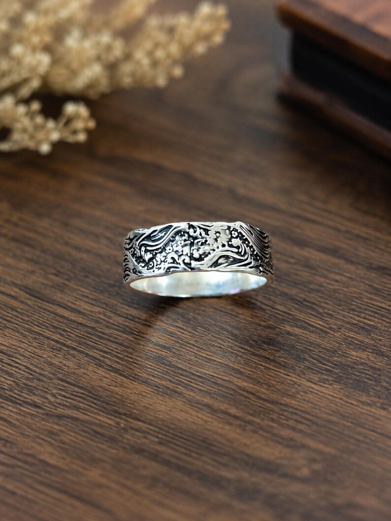 May include: A silver band ring featuring an intricate, raised relief pattern of swirling waves and small floral elements. The metal has a polished finish on the interior and a darkened, oxidized finish within the recessed areas of the design.