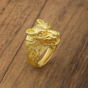 Könnte beinhalten: Ein goldfarbener Ring mit einem detaillierten Wolfskopf-Design. Der Ring hat eine strukturierte Oberfläche und ist vor einem Holz Hintergrund platziert. Die Merkmale des Wolfes sind klar definiert und zeigen die Handwerkskunst des Schmucks.