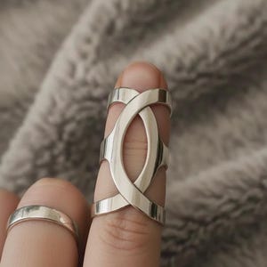 May include: A silver-colored ring with an intricate design worn on a finger. The ring features an open, oval-shaped center with intersecting bands. Another simple silver band ring is also visible.