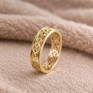 May include: A gold-colored ring with an intricate Celtic knot design. The ring features an openwork pattern, showcasing the detailed knotwork. The ring is set against a neutral, textured background.