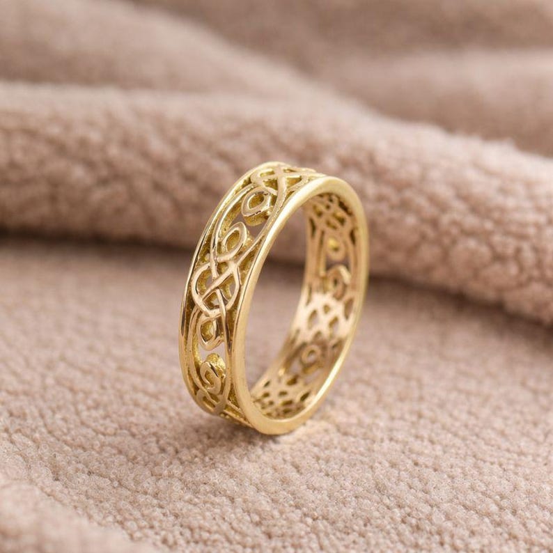 May include: A gold-colored ring with an intricate Celtic knot design. The ring features an openwork pattern, showcasing the detailed knotwork. The ring is set against a neutral, textured background.