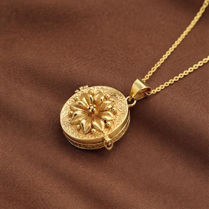 Peut inclure: Un collier avec un pendentif en forme de médaillon doré. Le médaillon rond présente une fleur en relief et des détails complexes. La chaîne est fine et le fermoir est visible.
