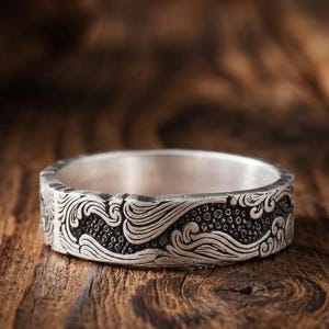 Op de afbeelding: Een zilveren ring met een gedetailleerd reliëfpatroon van wervelende golven en gestructureerde stippen. Het metaal heeft een gepolijste afwerking aan de binnenkant en een donker, geoxideerd contrast in de verdiepte delen van het golfpatroon.