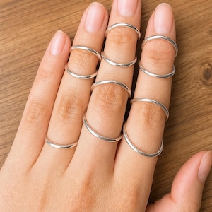 Könnte beinhalten: Eine Hand zeigt mehrere silberne Midi-Ringe. Die Ringe sind dünne Bänder aus poliertem Metall, die an den Fingern getragen werden. Die Ringe sind schlicht gestaltet und haben eine glatte, reflektierende Oberfläche. Der Hintergrund ist eine Holzoberfläche.