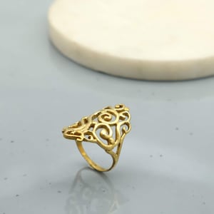 May include: A gold-coloured ring with an openwork design featuring swirling patterns. The ring has a triangular shape with a band for the finger. The ring is set against a light grey background.