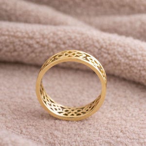 May include: A gold-colored ring with an openwork design. The ring features a repeating pattern of interconnected shapes, creating a delicate and intricate appearance. The ring is set against a soft, neutral-toned background.