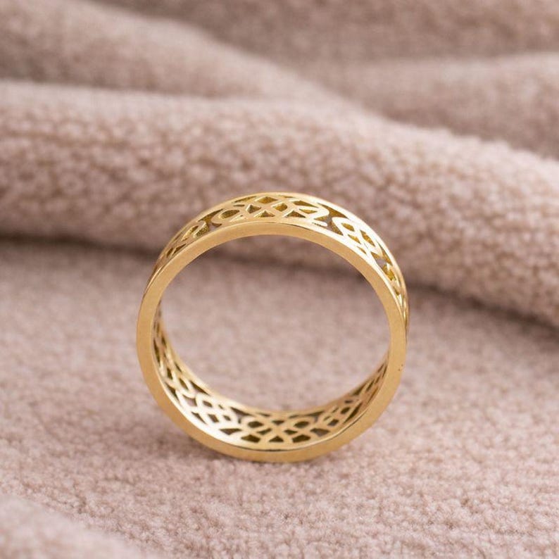 May include: A gold-colored ring with an openwork design. The ring features a repeating pattern of interconnected shapes, creating a delicate and intricate appearance. The ring is set against a soft, neutral-toned background.