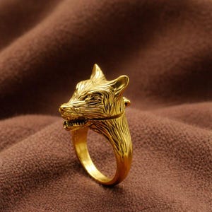 May include: A gold-colored ring featuring a detailed wolf head design. The ring's wolf head has intricate carved details, including the eyes, ears, and teeth. The ring is set against a brown background.