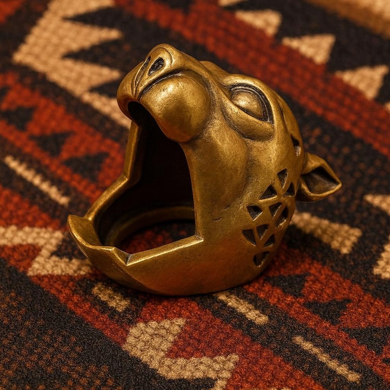 K&ouml;nnte beinhalten: Ein bronzefarbener Ring in Form eines br&uuml;llenden Jaguar-Kopfes. Der Ring weist detaillierte Gesichtsz&uuml;ge auf, darunter offene Kiefer und dreieckige Aussparungen an der Seite. Der Ring ist vor einem gemusterten Hintergrund platziert.