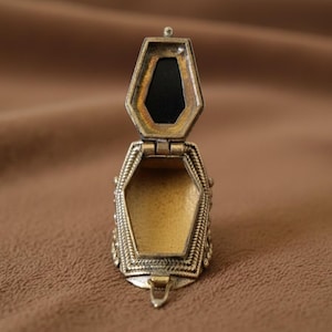 May include: An open, antique-style locket with a hinged lid. The locket is made of a gold-coloured metal with intricate detailing around the edges. The interior is a light gold colour, and the lid has a black, rectangular insert.