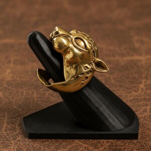 K&ouml;nnte beinhalten: Ein goldfarbener Ring mit einem detaillierten Jaguar-Kopf-Design. Der Ring wird auf einem schwarzen, abgewinkelten St&auml;nder pr&auml;sentiert, der seine komplizierten Merkmale hervorhebt. Das Gesicht des Jaguars ist mit sichtbaren Augen, Ohren und einer strukturierten M&auml;hne geformt.