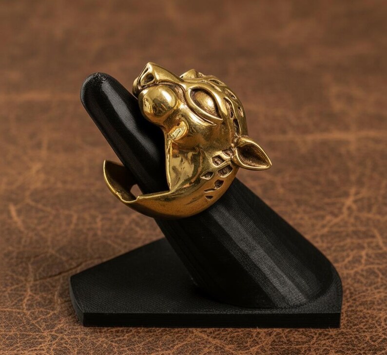 K&ouml;nnte beinhalten: Ein goldfarbener Ring mit einem detaillierten Jaguar-Kopf-Design. Der Ring wird auf einem schwarzen, abgewinkelten St&auml;nder pr&auml;sentiert, der seine komplizierten Merkmale hervorhebt. Das Gesicht des Jaguars ist mit sichtbaren Augen, Ohren und einer strukturierten M&auml;hne geformt.