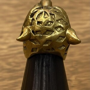 K&ouml;nnte beinhalten: Ein detaillierter, goldfarbener Ring mit einem strukturierten, durchbrochenen Design. Der Ring hat zwei hornartige Vorspr&uuml;nge auf beiden Seiten und wird auf einem schwarzen St&auml;nder pr&auml;sentiert. Das filigrane Muster und der metallische Glanz des Rings sind deutlich sichtbar.