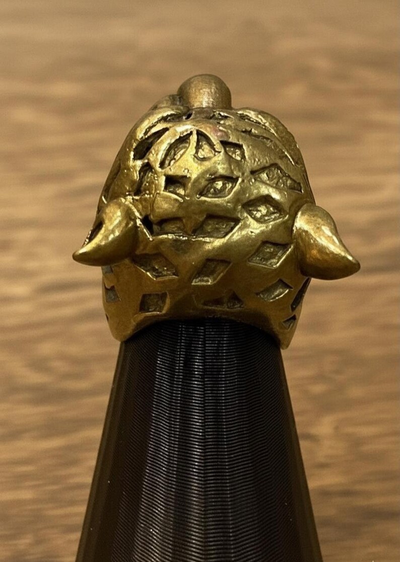 K&ouml;nnte beinhalten: Ein detaillierter, goldfarbener Ring mit einem strukturierten, durchbrochenen Design. Der Ring hat zwei hornartige Vorspr&uuml;nge auf beiden Seiten und wird auf einem schwarzen St&auml;nder pr&auml;sentiert. Das filigrane Muster und der metallische Glanz des Rings sind deutlich sichtbar.