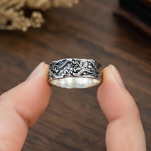 May include: A silver ring featuring an intricate, embossed pattern of swirling waves and small floral elements. The metal has a darkened, oxidized finish within the recessed areas of the design, creating a high-contrast look against the polished silver.