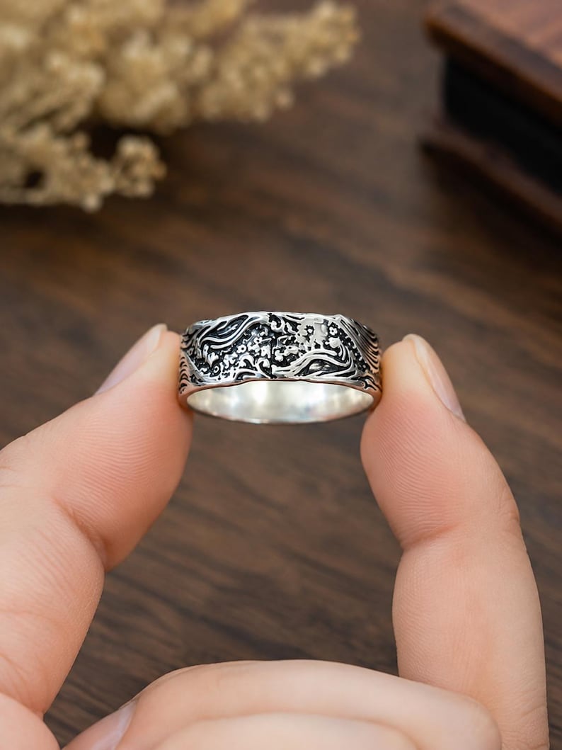 May include: A silver ring featuring an intricate, embossed pattern of swirling waves and small floral elements. The metal has a darkened, oxidized finish within the recessed areas of the design, creating a high-contrast look against the polished silver.