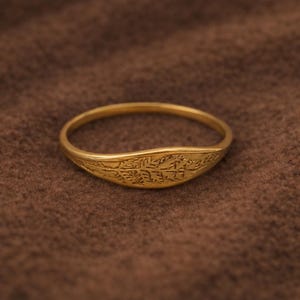 May include: A gold ring featuring an engraved design. The ring has a smooth, rounded band and a curved face with detailed patterns. The ring is displayed against a brown backdrop. The ring's design is intricate.