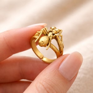 Pode incluir: Um anel dourado com um design detalhado de uma formiga. O anel tem uma banda ajustável e a formiga é esculpida com detalhes intrincados, incluindo pernas, antenas e um corpo segmentado. O anel é segurado por uma pessoa.