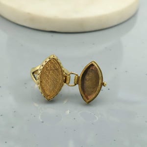 Peut inclure: Une bague locket dorée avec un compartiment ovale qui s'ouvre. La bague a une bordure texturée autour du compartiment. La bague est sur une surface gris clair.