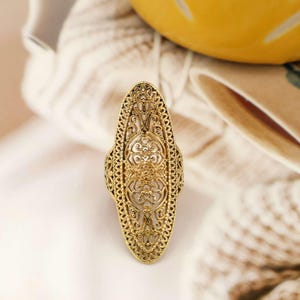Peut inclure: Une bague filigrane dorée de forme ovale allongée. La bague présente des motifs floraux et de volutes complexes. Ce bijou est une pièce maîtresse, parfaite pour ajouter une touche d'élégance vintage à n'importe quelle tenue.