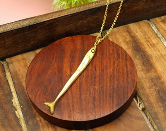 May include: A gold-colored fish pendant necklace. The pendant has a textured surface and a small eye detail. The necklace is displayed on a round, dark wooden surface, with a wooden tray in the background. The fish pendant is suspended from a delicate chain.