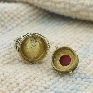 May include: A detailed, open brass ring with an ornate design. The ring has a large, open setting. Beside it is a matching brass component with a red stone. The items are on a textured, off-white surface.
