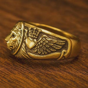 Può includere: Un anello color oro con una testa di leone dettagliata su un lato e un cuore con ali sull'altro. L'anello presenta un design intricato con un'estetica vintage o antica. L'anello è probabilmente in ottone o metallo simile.
