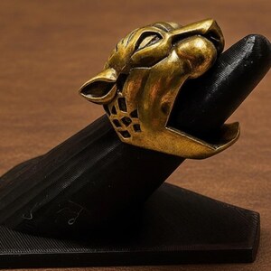 K&ouml;nnte beinhalten: Ein goldfarbener Ring mit einem br&uuml;llenden Jaguar-Kopf-Design. Der Ring wird auf einem schwarzen Ringhalter pr&auml;sentiert. Das Maul des Jaguars ist ge&ouml;ffnet und zeigt Z&auml;hne. Der Ring hat ein detailliertes, strukturiertes Aussehen.