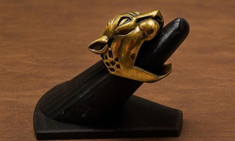K&ouml;nnte beinhalten: Ein goldfarbener Ring mit einem br&uuml;llenden Jaguar-Kopf-Design. Der Ring wird auf einem schwarzen Ringhalter pr&auml;sentiert. Das Maul des Jaguars ist ge&ouml;ffnet und zeigt Z&auml;hne. Der Ring hat ein detailliertes, strukturiertes Aussehen.