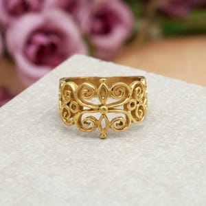 Può includere: Un anello dorato con un design traforato e ornato. L'anello presenta un motivo ripetuto di volute e forme floreali, creando un aspetto delicato e dettagliato. L'anello è probabilmente in metallo e viene presentato su una superficie neutra.