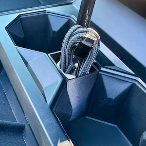 May include: Black plastic car console organizer with multiple compartments. A black pen and a gray and black braided USB cable are in the organizer. The console is in a vehicle.