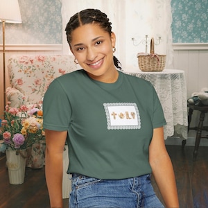 May include: A green t-shirt with a small embroidered patch featuring the word "HOLY" and religious symbols. The patch is framed by a white and gray lace-like border. The shirt is paired with blue jeans.