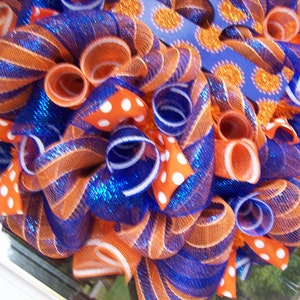May include: A wreath made of orange and blue mesh with white polka dot ribbon. The wreath is decorated with a blue ribbon with orange glitter dots.