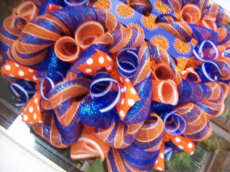May include: A wreath made of orange and blue mesh with white polka dot ribbon. The wreath is decorated with a blue ribbon with orange glitter dots.