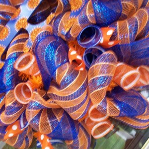 May include: A wreath made of orange and blue mesh ribbon with white polka dots. The wreath is decorated with a blue and orange glitter bow.